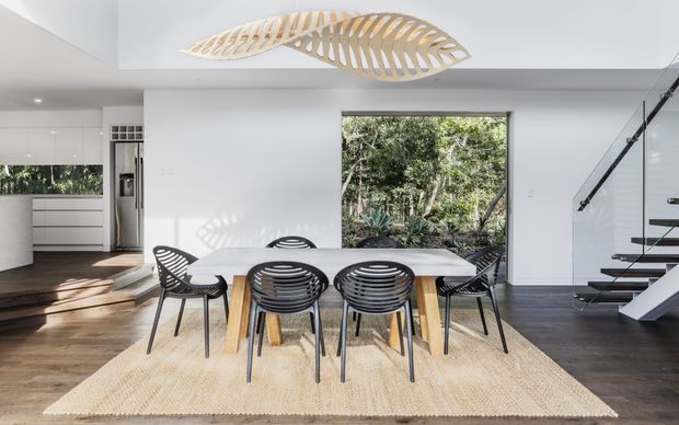 Bright open-plan dining area with a white rectangular dining table and black woven chairs arranged on a natural beige rug. The space connects to a sleek white kitchen at left and a floating staircase with glass balustrade at right. Dark timber flooring, white walls and cabinetry, large garden-facing glazing, and a sculptural timber ceiling light create a clean contemporary interior.