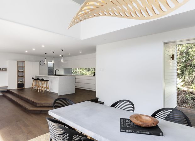 Bright open-plan kitchen and dining area with white walls and cabinetry, a compact island with timber bar stools, dark timber flooring, and a large pale dining table with sculptural black chairs. A distinctive suspended timber feature light hangs above the dining zone, while large windows and a glazed door bring in views of greenery.