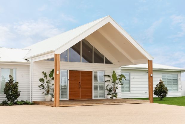 Single-storey white-clad house exterior with a prominent gabled entry porch. The facade features horizontal weatherboard cladding, a pale metal pitched roof, tall timber porch posts, a timber deck, large timber double entry doors, louvred sidelights, and a triangular glazed gable above the entrance. Minimal landscaping and a broad pale driveway are visible in front.