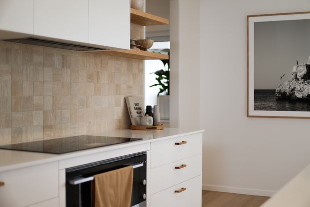 A compact, softly styled kitchen with white flat-panel cabinetry, pale timber open shelving, a light beige textured tile backsplash, and a dark induction cooktop above a built-in oven. Timber handles, neutral walls, timber flooring, and a framed monochrome artwork create a warm, refined contemporary interior.