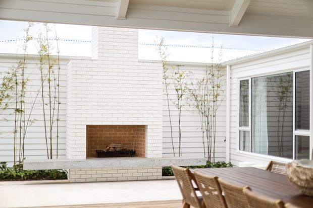 Bright covered outdoor dining and entertaining area with a built-in white-painted brick fireplace, horizontal weatherboard walls, white-framed glazing, pale paving, and a timber dining table with woven chairs. Slender bamboo planting softens the courtyard-like setting.