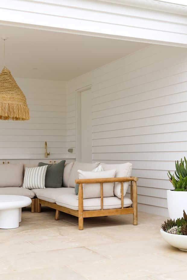 A bright covered outdoor lounge with white horizontal weatherboard walls, pale tiled flooring, and a relaxed sofa arrangement. The seating has light upholstered cushions and timber framing, complemented by a woven pendant light, white round coffee table, and potted succulents. The space has a breezy coastal feel with warm natural accents.