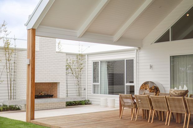 Covered outdoor dining terrace adjoining a white weatherboard-clad home, with a pitched gabled roof and exposed white-painted rafters. The space features a timber deck, large woven dining table and chairs, white brick walls, and a built-in outdoor fireplace opening onto a minimalist courtyard.