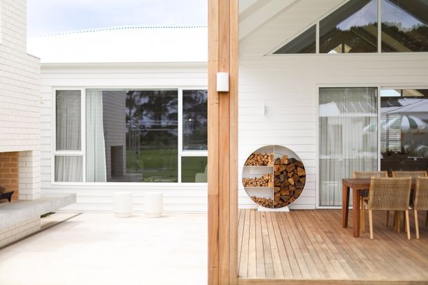 Bright contemporary courtyard-style outdoor living area with white horizontal brick walls, timber decking, a substantial timber post, large glazed doors and windows, and a built-in circular firewood storage feature. The space includes an outdoor dining setting and opens around a pale paved courtyard.