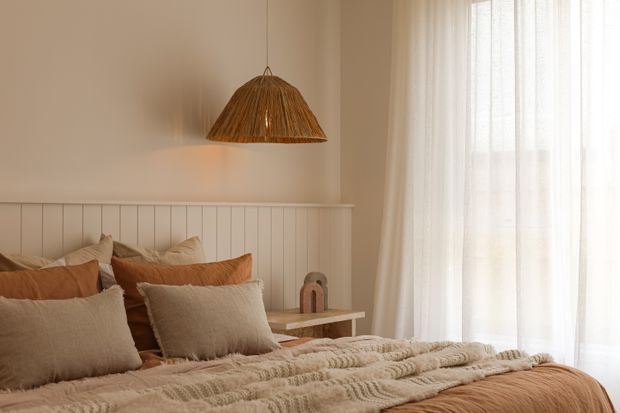 A warm, softly lit bedroom with a low bed dressed in layered beige, cream and terracotta textiles. The bed sits against a white vertical-panel headboard, with a woven pendant light above and sheer floor-to-ceiling curtains filtering daylight from a large window or glazed door. A small timber bedside table and minimal decorative object complete the calm, natural interior.