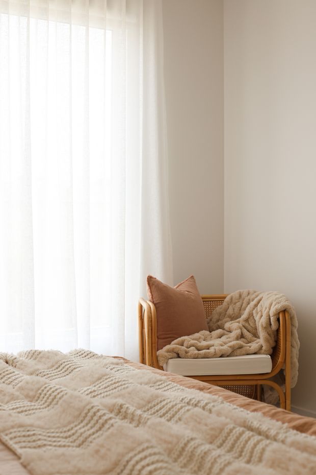 A softly lit bedroom corner with a bed covered in a textured cream blanket, a rattan-framed accent chair with a warm beige cushion and throw, sheer white curtains, and neutral beige walls. The scene has a cozy, calm, natural feel.