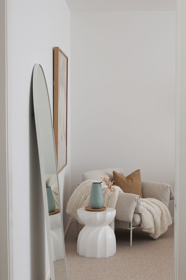 A softly styled, neutral-toned living room corner featuring a pale upholstered lounge chair, textured cream throw, tan cushion, sculptural white side table, and muted teal vase. A tall rounded floor mirror leans against the wall beside framed artwork, creating a calm and refined interior vignette.