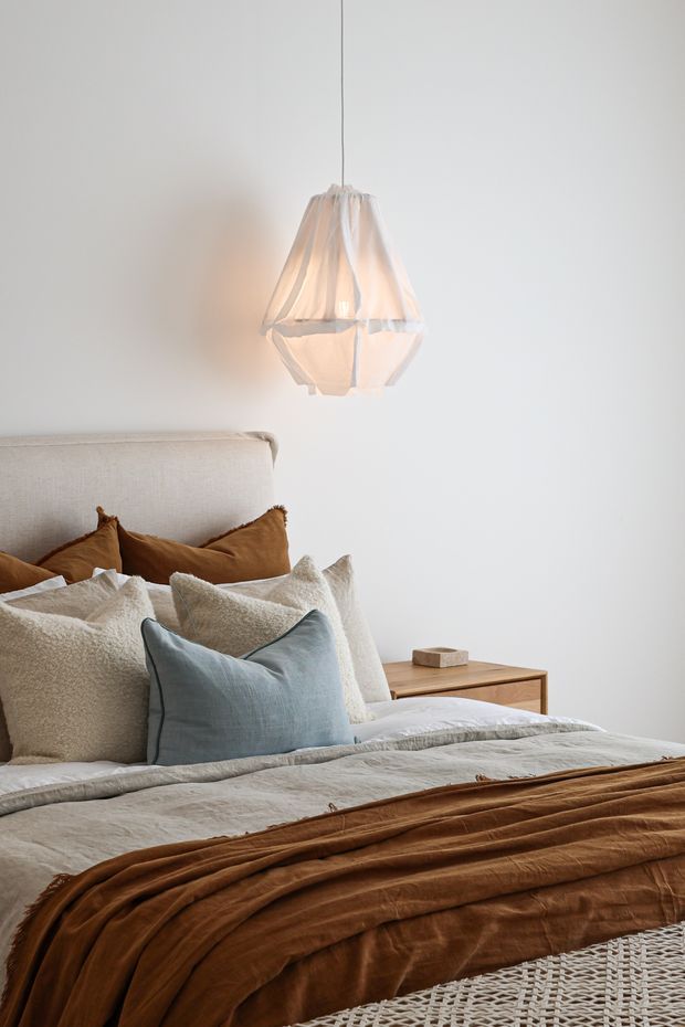 A softly lit, calm bedroom featuring a neutral upholstered bedhead, layered cream and textured beige pillows, a muted blue accent cushion, pale bedding, and a rich brown throw. A simple timber bedside table and sculptural fabric pendant light complement the minimal white room.