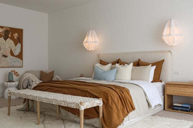 A warmly styled bedroom with a low upholstered bed, layered white and rust-brown bedding, pale blue and cream cushions, and a woven fringed bench at the foot. Two sculptural pendant lights hang above the bed, while a simple timber bedside table, boucle chair, soft rug, and framed artwork complete the calm, contemporary interior.