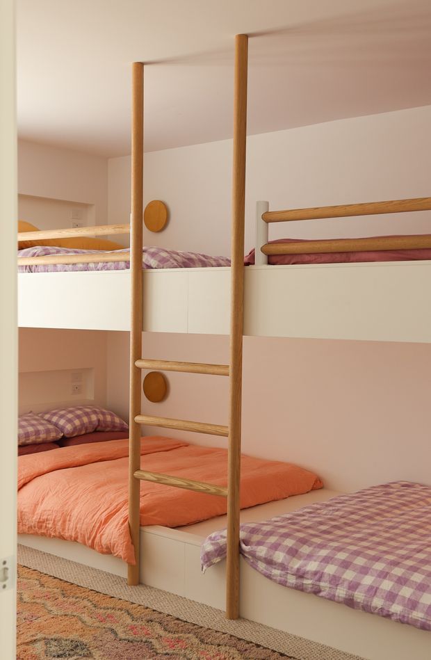 A compact children’s bunk bedroom with custom built-in white bunk beds, a central timber ladder, and rounded timber guard rails. The bunks are dressed with coral, pink, and purple gingham bedding, while a patterned rug adds warmth to the lower floor. Soft cream-pink walls and warm timber details create a playful, cozy atmosphere.