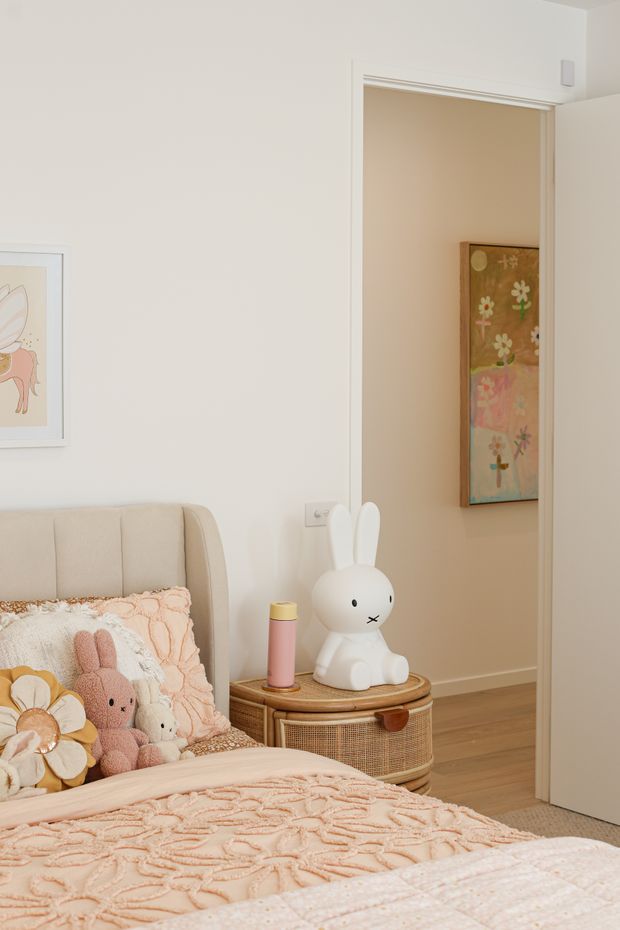 A softly styled children's bedroom in warm neutral and blush tones. The room features a partially visible upholstered bed with textured pink bedding and plush toys, a woven wicker bedside cabinet with a white rabbit lamp, pale walls, framed artwork, and an open doorway leading to an adjacent space.