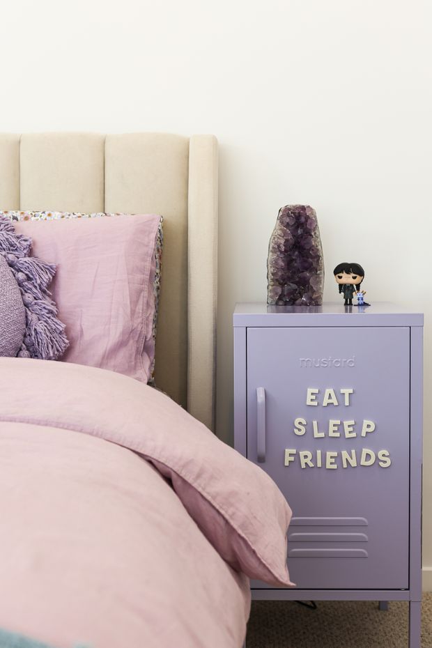 A close-up view of a softly styled bedroom corner, featuring a blush-pink bed with a cream upholstered headboard and lavender bedding. A lavender bedside cabinet with raised lettering reading “EAT SLEEP FRIENDS” sits beside the bed, topped with an amethyst geode and small figurines. The room has a playful, pastel-toned aesthetic.