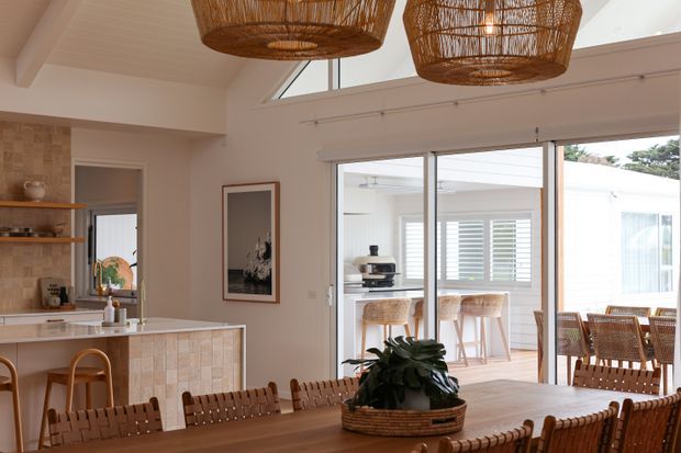 Warm, light-filled open-plan dining and kitchen interior with a large timber dining table and woven dining chairs in the foreground. The kitchen features pale cabinetry, a tiled island face, open timber shelving and stone-look benchtops. Large sliding glass doors connect to a bright outdoor entertaining area with bar seating, while woven pendant lights and rattan furniture add a relaxed coastal character.