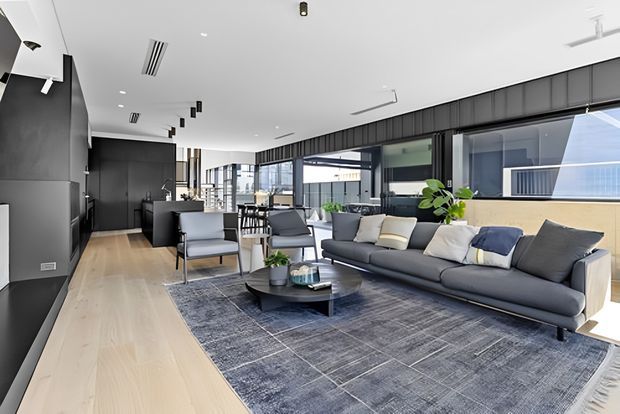 Bright contemporary open-plan living area with a large charcoal sectional sofa, lounge chairs, round coffee table, and oversized grey patterned rug. Pale timber flooring contrasts with dark built-in cabinetry and exposed dark-framed glazing. The room extends toward a dining area and kitchen, with abundant natural light and indoor plants.