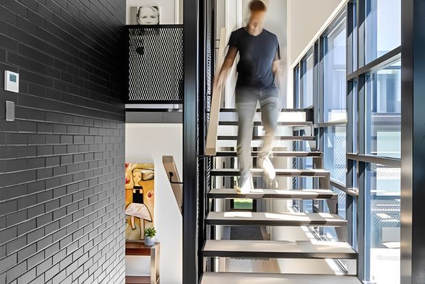 Bright contemporary stairway with open-riser floating timber steps and dark metal supports, positioned beside a full-height glazed wall. A dark charcoal brick feature wall contrasts with white walls and abundant natural light. The stairs connect to an upper level, with glimpses of a furnished room through the adjacent opening.