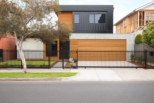 Street-facing view of a contemporary two-storey residence with a flat roof, dark vertical cladding, warm timber wall and garage-door finishes, a concrete garage surround, and a black metal front fence and gate. The property includes a driveway, landscaped front garden, and mature street tree.