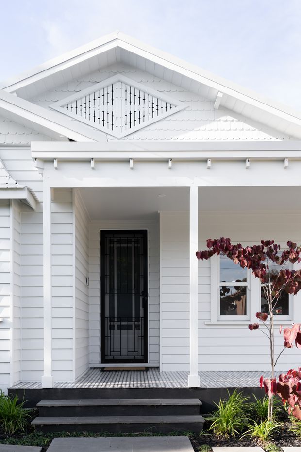 Front elevation of a white weatherboard home featuring a covered entry porch, decorative gable infill, black security screen door, white posts and trim, patterned tiled porch floor, dark steps, and simple landscaped planting.