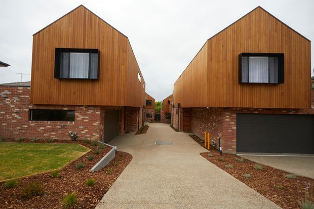 Exterior view of a pair of contemporary gable-fronted dwellings arranged along a shared concrete driveway. The buildings combine warm vertical timber cladding on the upper façades with exposed red brick lower levels, black-framed windows, and an integrated dark garage door. Low-maintenance landscaped garden beds and a central pedestrian path are visible.