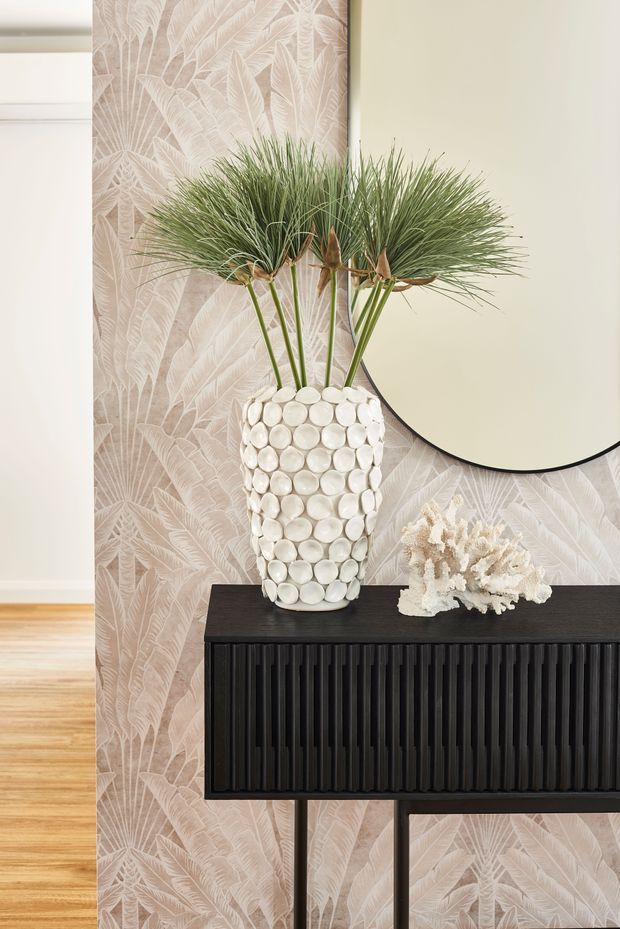 A refined interior vignette featuring a slim black console with vertically fluted detailing, positioned against pale botanical-patterned wallpaper. A white textured vase filled with tall ornamental grasses and a coral-like decorative object sit on the console, while a large round wall mirror is partially visible above. Timber flooring and light neutral finishes create a warm, elegant atmosphere.