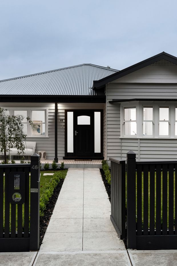 Front exterior view of a compact single-storey house with grey horizontal weatherboard cladding, a corrugated metal gable roof, black-framed entry door and windows, and a black picket fence enclosing a landscaped front path. The façade has a clean contemporary renovation or bungalow character.