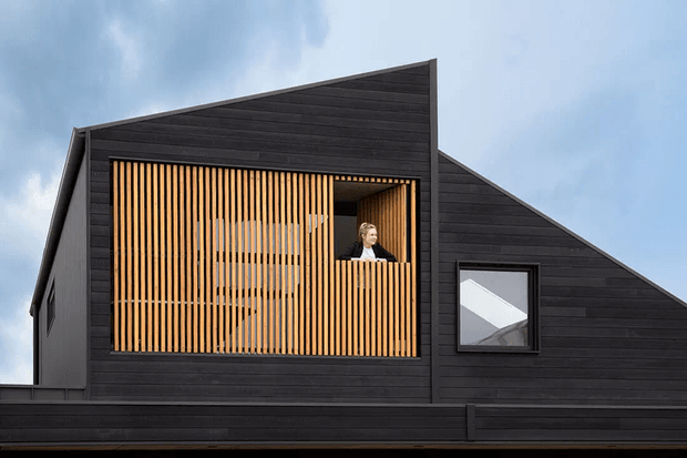 Exterior view of a dark-clad, angular gable-roofed building with a prominent upper-level balcony or recessed opening screened by closely spaced vertical timber battens. A person is visible behind the screen, and a small square window sits in the side wall. The facade has a strong geometric composition and contrasting warm timber screening.