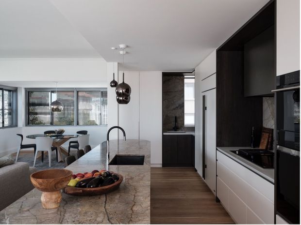 Open-plan contemporary kitchen and dining area with a large veined stone island, dark and white built-in cabinetry, integrated appliances, timber flooring, pendant lights, and a dining table beside horizontal blinds and glazing. A partial living area is visible in the foreground.