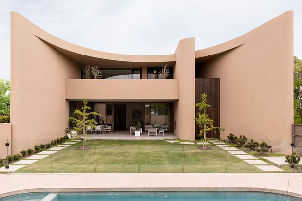Front exterior view of a sculptural two-storey residence with sweeping curved terracotta-toned walls and roofline, a recessed upper balcony, dark timber screening, extensive glazing, and an open ground-floor entertaining area. A formal lawn with stepping-stone paths leads toward the house, with a swimming pool visible in the foreground.