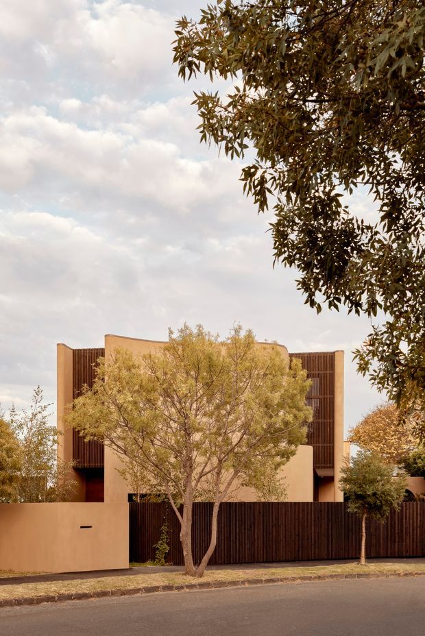 Street-facing view of a contemporary multi-level residence behind a low rendered wall and dark vertical timber screening. The facade combines warm earth-toned plaster volumes with extensive vertical battens, partially obscured by mature street trees and landscaping. The image has a warm, softly lit suburban architectural photography style.