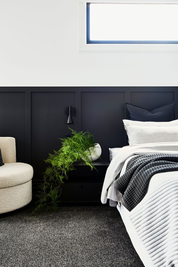 A contemporary bedroom featuring a dark charcoal-black paneled feature wall and matching low bedside furniture. The bed is dressed in layered white bedding with a textured dark grey throw and navy cushions. A cream boucle armchair, sculptural wall light, potted fern, and high horizontal window complete the refined monochromatic scheme. Dark carpet covers the floor.