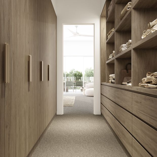 A narrow walk-in wardrobe or dressing room with full-height wood-grain cabinetry on both sides. One side has tall closed wardrobes with long vertical handles, while the other has open shelving, folded clothing, and wide drawers. Beige carpet and warm timber finishes lead toward a bright adjoining living area with large glazing.