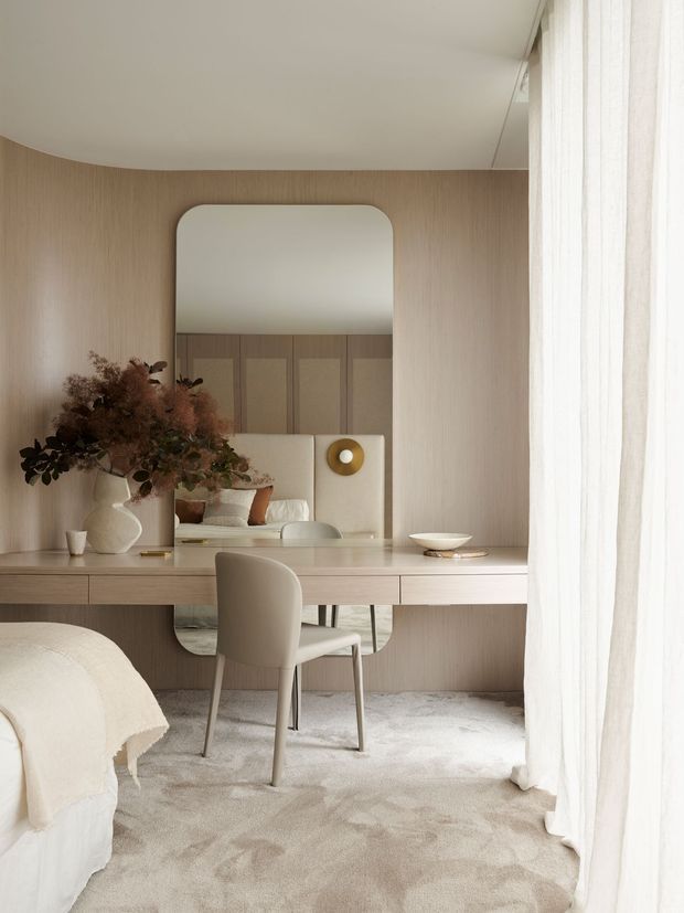 A serene, softly lit bedroom interior featuring a built-in timber-toned dressing desk spanning the wall, a rounded-rectangle mirror, upholstered chair, neutral carpet, sheer curtains, and a partially visible bed. The space uses warm beige and off-white tones with subtle organic decor and minimalist built-in joinery.