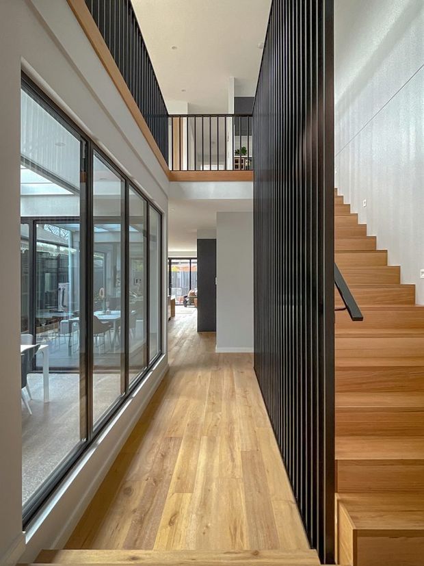 A bright contemporary interior hallway beside a timber staircase. Full-height glazing on the left looks into a light-filled room, while a tall black vertical slatted screen separates the stair from the circulation space. The hallway has warm timber flooring, white walls and ceiling, black-framed glazing, and an upper-level landing with matching black balustrades.