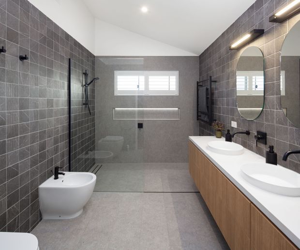 Contemporary bathroom with a spacious walk-in shower, wall-hung toilet and bidet, double vanity, oval mirrors, and dark grey tiled walls. The room has a restrained neutral palette with warm timber cabinetry, white sanitaryware, and integrated modern lighting.