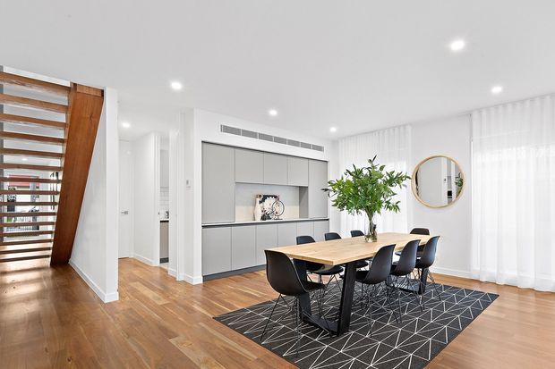 Bright open-plan dining area with a contemporary built-in grey media or storage wall, timber flooring, a black dining table and chairs, and a geometric black-and-white rug. Floor-to-ceiling sheer curtains, recessed ceiling lighting, a round wall mirror, indoor greenery, and a timber louvred stair or screen feature are visible.