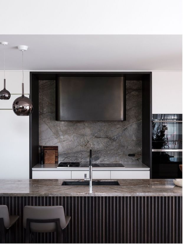 A sophisticated contemporary kitchen viewed from the island, featuring a dramatic grey veined stone splashback and benchtops, black-framed recessed cabinetry, integrated dark rangehood, induction cooktop, built-in oven, and a fluted dark island front. Chrome pendant lights and a minimalist black tap provide reflective accents, with seating for dining at the island.