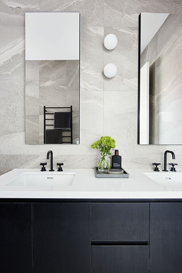 Contemporary double vanity bathroom with a long white benchtop, two integrated rectangular basins, matte black wall-mounted and deck-mounted tapware, dark vertically ribbed vanity cabinetry, large frameless mirrors, and pale grey veined stone-look wall tiles. Round wall lights and a towel ladder add simple functional detailing.