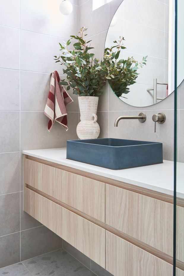 A bright contemporary bathroom featuring a floating light timber vanity with integrated drawers, a white countertop, and a deep blue vessel basin. Large pale grey wall tiles and terrazzo-style floor tiles create a clean backdrop, while brushed metal wall-mounted tapware, a rounded mirror, and a leafy decorative arrangement add visual interest.