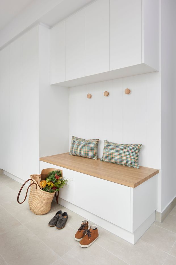 Bright minimalist entryway featuring full-height white built-in cabinetry, a timber-topped bench seat with patterned cushions, simple wooden wall hooks, and pale large-format tiled flooring. A woven basket and shoes sit on the floor, reinforcing the practical mudroom-style entrance.