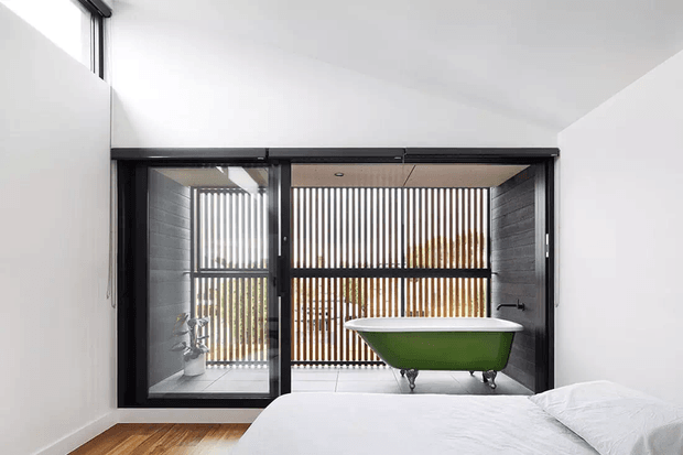 A bright contemporary bedroom with a freestanding green bathtub visible through large black-framed glazed doors. The room features white walls and bedding, timber flooring, and a dramatic vertical timber screen filtering light behind the bath. A small adjacent washroom or bathing zone is visible to the left, with a plant and additional glazing.
