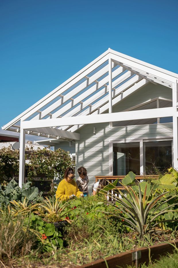 Exterior view of a light-coloured weatherboard house with a prominent white timber pergola or veranda structure and a steep gable roof with glazed or translucent slatted screening. The home opens onto a lush planted garden with raised beds, and several people are present in the foreground. Bright blue sky and strong sunlight create a fresh residential outdoor setting.