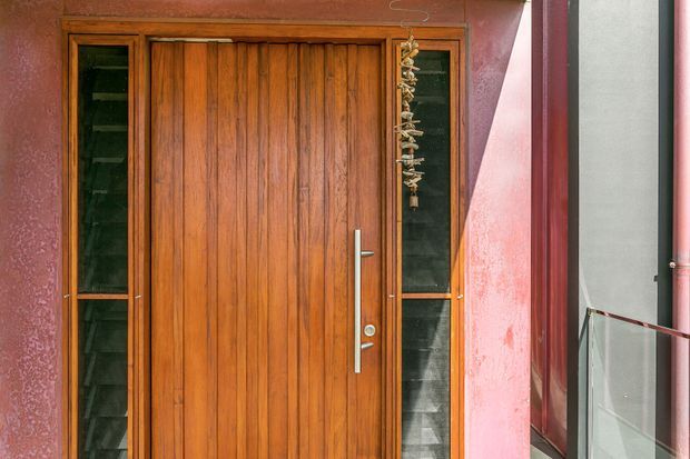 Close-up view of a residential entry door featuring a tall warm-toned timber door with vertical grain, narrow glazed sidelights, and a long brushed-metal pull handle. The entry is framed by textured pink-red plaster walls and dark trim, with a glass balustrade visible to the right.