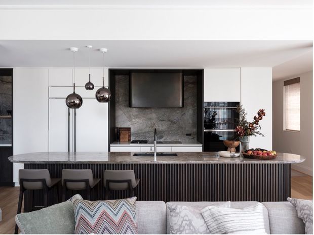 Contemporary open-plan kitchen viewed from the living area, featuring a long stone island with integrated sink and seating for three, dark vertically fluted cabinetry, white full-height cabinetry, a dark stone splashback and television niche, and three spherical pendant lights. A sofa occupies the foreground, while timber flooring and a window are visible to the right.