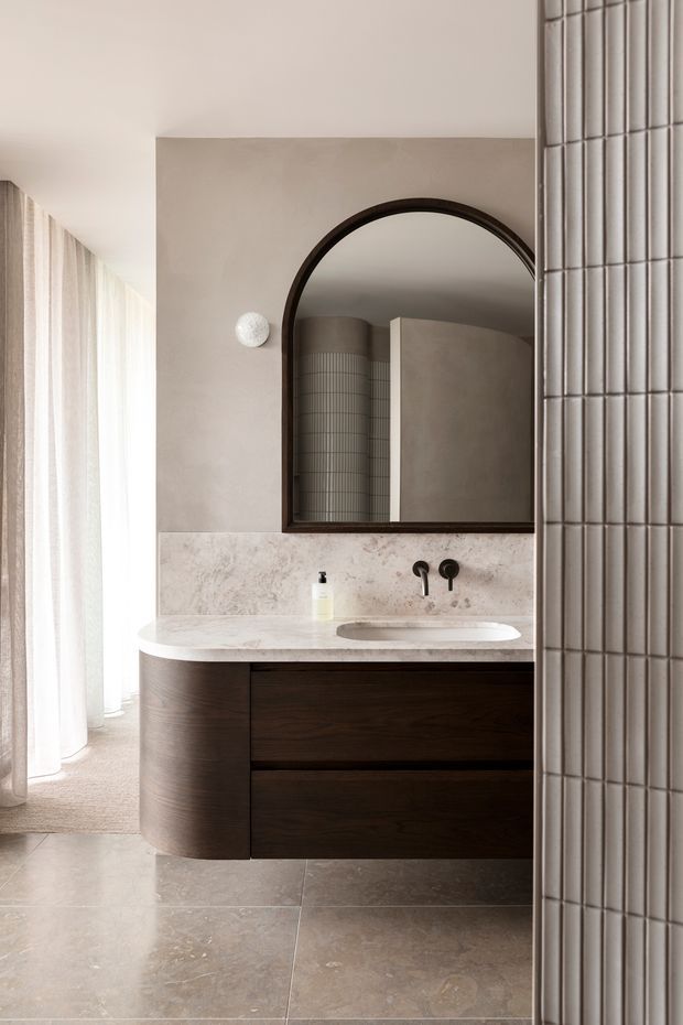 A refined contemporary bathroom featuring a floating dark timber vanity with a curved end, a pale stone benchtop and integrated basin, black wall-mounted tapware, and a large arched dark-framed mirror. Warm beige plaster walls, light stone-look floor tiles, sheer curtains, and a curved tiled feature wall create a calm, sculptural interior.