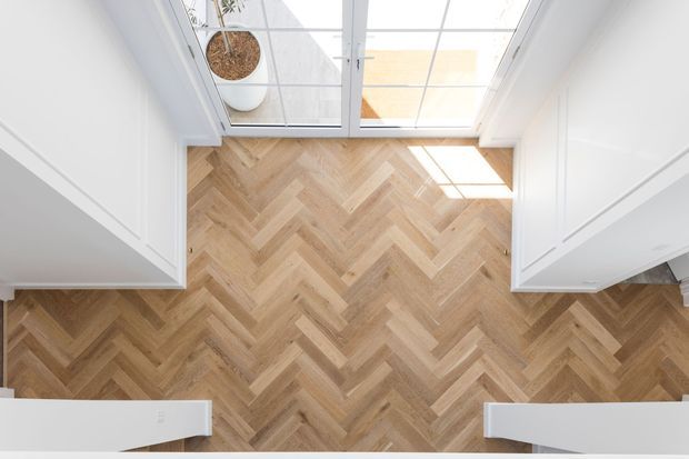 Top-down view of a bright interior hallway or entry space with warm herringbone timber flooring, white paneled walls and cabinetry, and a glazed roof or skylight bringing in abundant natural light. A potted plant is visible near the glazing.