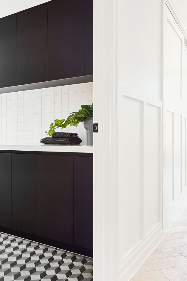 A high-contrast interior view looking from a bright white paneled hallway into a compact kitchen. The kitchen features matte near-black cabinetry, a white tiled splashback, a white benchtop, dark patterned geometric floor tiles, and a small potted plant. The hallway has white decorative wall panelling, white trim, and pale timber flooring.