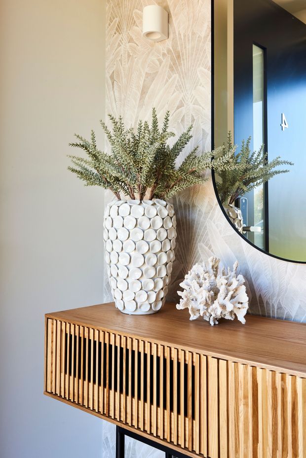 Close-up interior view of a refined entryway or hallway vignette featuring a wall-mounted slatted timber console, a large round black-framed mirror, botanical patterned wallpaper, and a decorative white textured vase with greenery. The palette is warm timber, white, cream, and black, with layered natural textures and contemporary styling.