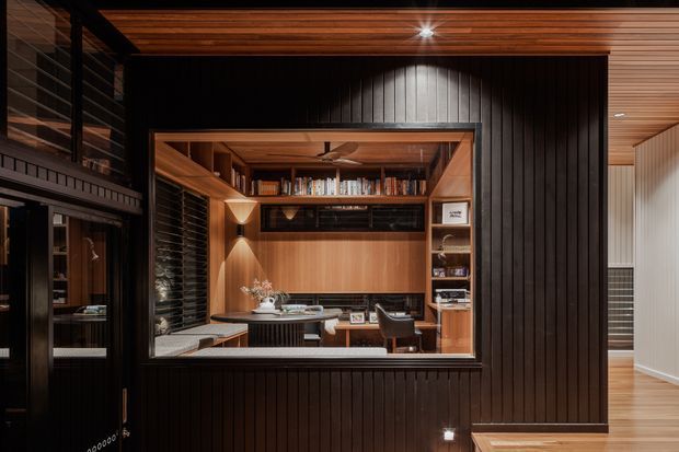 A dark, refined home office or study viewed through a large internal opening, with extensive warm timber joinery, built-in bookshelves, desk workspace, and concealed storage. The surrounding room features black vertical wall panelling, a timber slatted ceiling, and integrated lighting, creating a sophisticated contemporary interior.