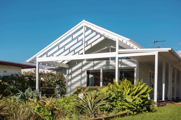 Exterior view of a light-coloured single-storey house with a prominent open gable roof structure, covered veranda or patio, extensive glazing, and white weatherboard-style cladding. The home is surrounded by lush garden planting under a clear blue sky.