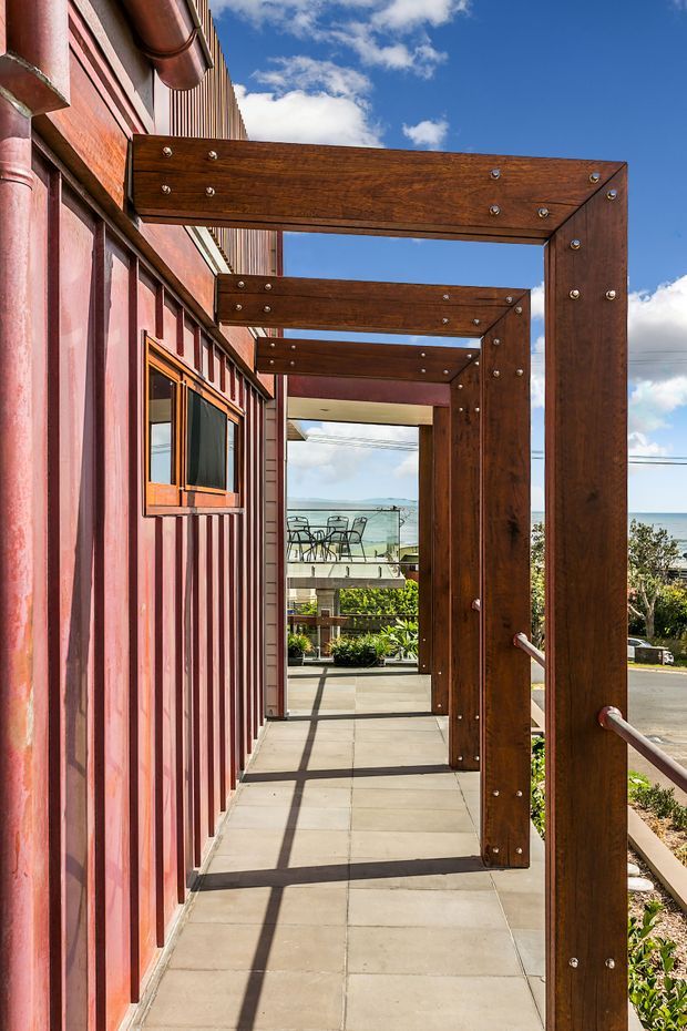 A covered exterior walkway alongside a red corrugated-clad building, framed by repeated dark timber portal structures with exposed metal fixings. Large concrete pavers lead toward a glazed outdoor area and a visible coastal horizon beyond. Bright daylight creates strong linear shadows across the path.