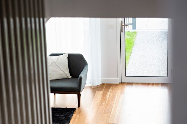 A bright, compact interior entry area with warm timber flooring, white walls, sheer curtains, and a glazed external door. A dark upholstered armchair with a light cushion sits beside a black rug, while vertical timber screening or slatted joinery partially frames the left side of the view.